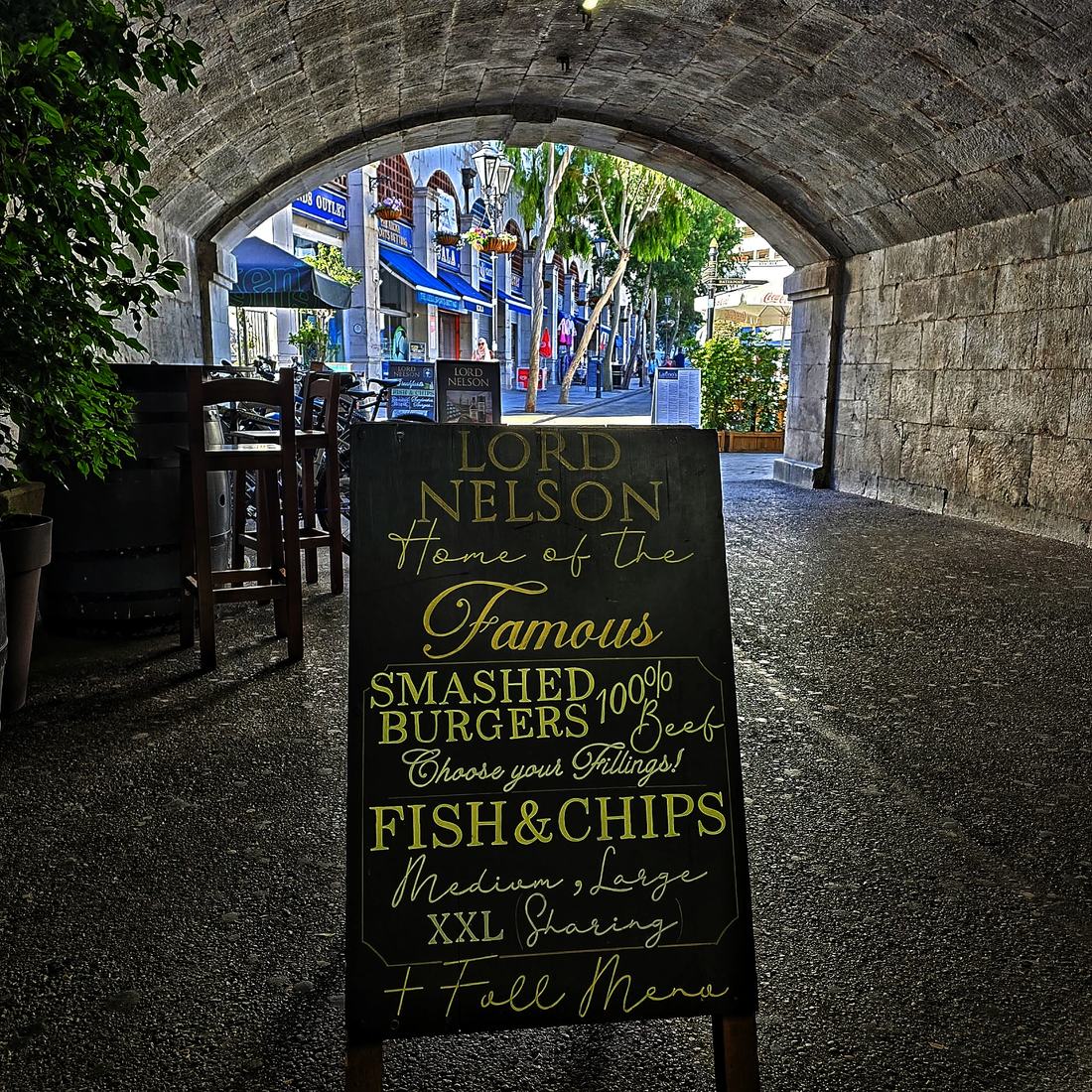 The Lord Nelson A-board under the Casemates arch in Gibraltar, reading “Home of the Famous Smashed Burgers and Fish & Chips — Medium, Large, XXL Sharing”